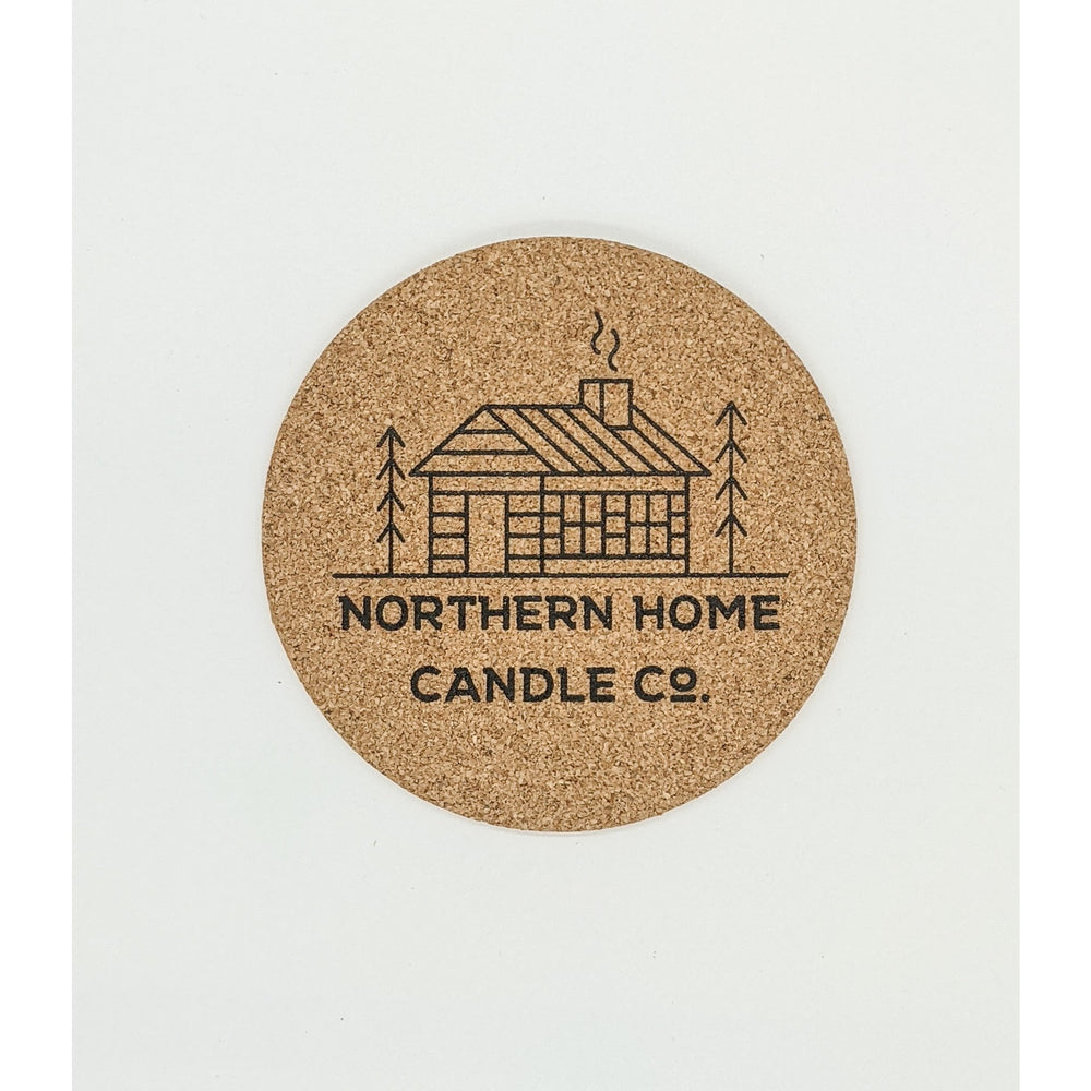 4 inch wide cork coaster with printed black image of a cabin and trees with the words northern home candle co underneath.