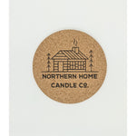 4 inch wide cork coaster with printed black image of a cabin and trees with the words northern home candle co underneath.