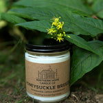 Candle jar labeled 'Honeysuckle Breeze' from Northern Home Candle Co. on a natural background with green leaves.