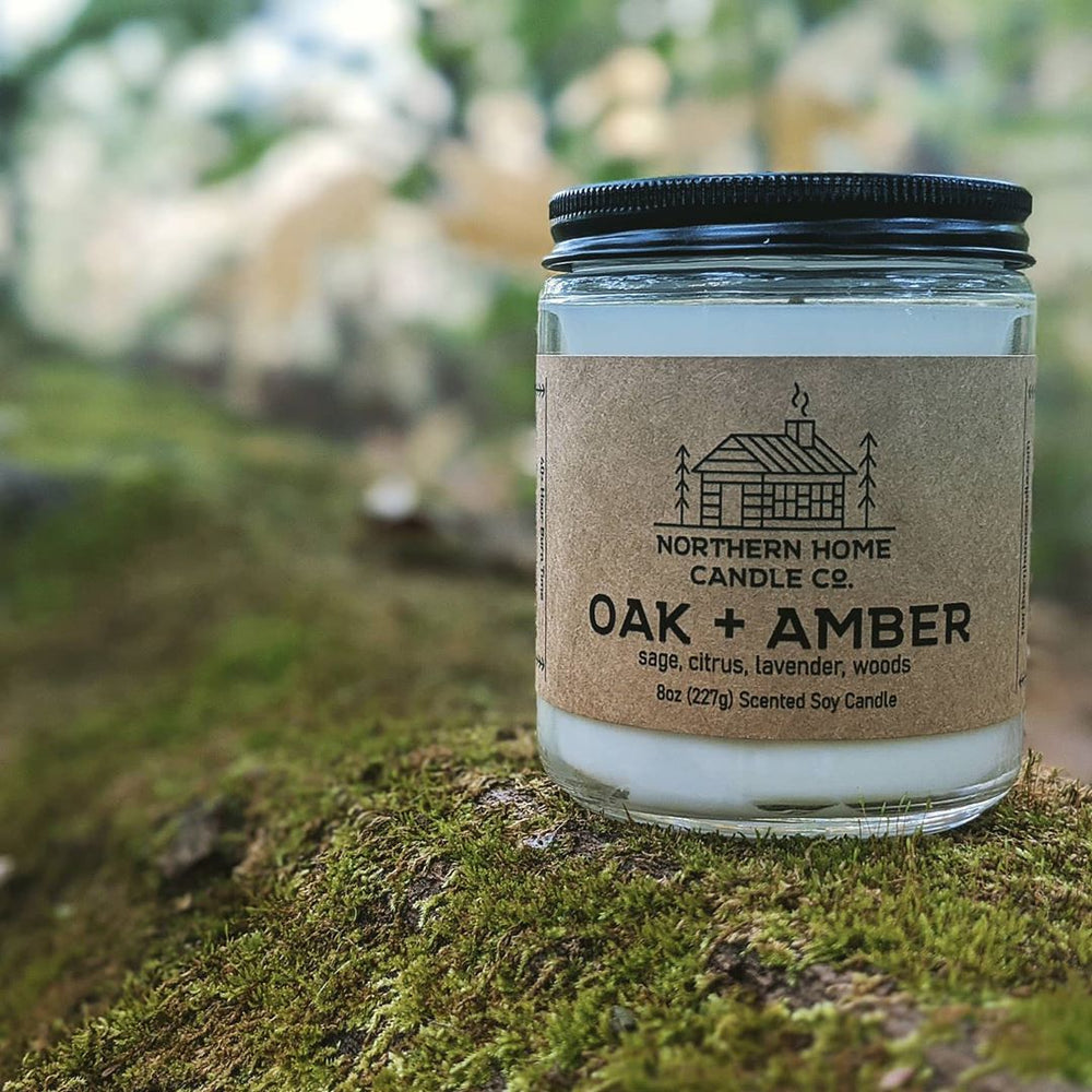 8 ounce soy candle sitting on a mossy log. The label is brown and reads northern home candle company and oak and amber. 