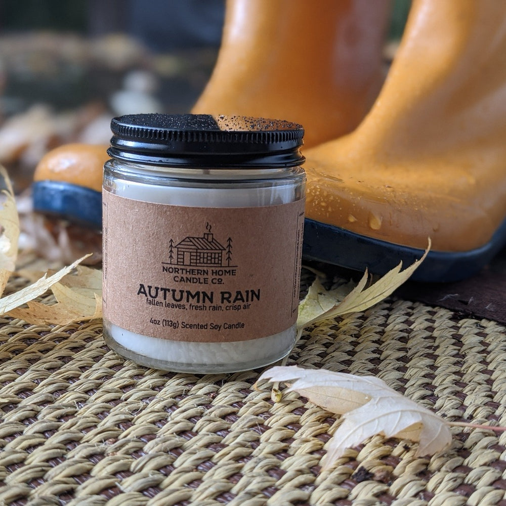 Soy candle with brown label that reads northern home candle company and autumn rain glazed with rain water and rain boots in the background.